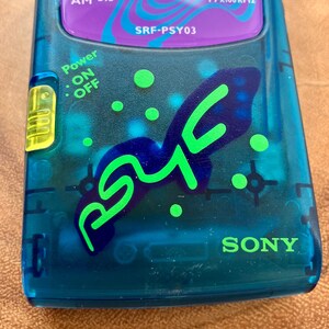 May include: A close-up of a vintage Sony SRF-PSY03 AM/FM radio. The translucent teal plastic case features a blue and green graphic design with the word "PSYc" and the Sony logo. The radio has a purple and teal top panel with the text "AM 530" and "FM x100kHz".