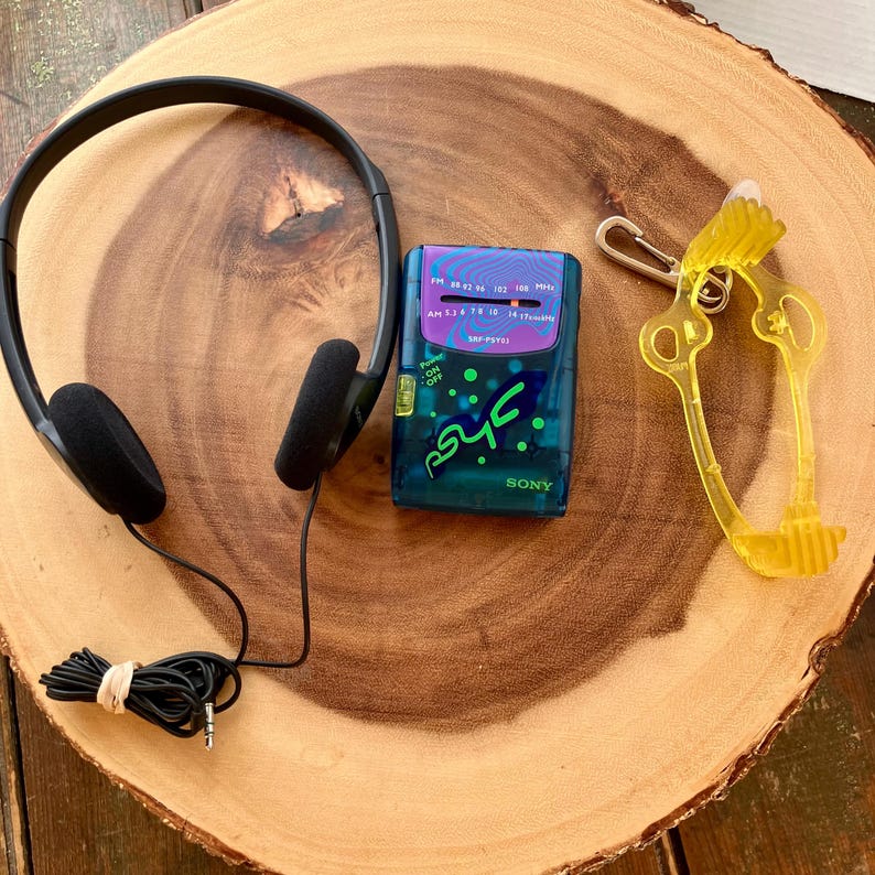 May include: A black over-the-ear headphones, a teal Sony SRF-FS01 radio, and a yellow key-shaped keychain are arranged on a wooden surface. The radio has a purple and blue design with the word "Sony" on the bottom. The headphones' cord is bundled.