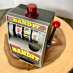 May include: A black and silver Jackpot Bandit slot machine toy featuring a red top light and a red side lever. The front displays the word BANDIT in yellow letters, three spinning reels with symbols like 7s and bars, and a coin return tray.