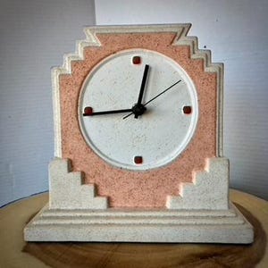 May include: A mantel clock with a stepped, geometric frame in speckled off-white and peach. The circular clock face is white with a speckled texture, featuring black hour and minute hands, a thin second hand, and four small red square markers.