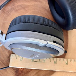 May include: Over-ear headphones with a silver and gray frame and black ear pads. The headphones have the brand name "SONY" printed on the ear cup. A wooden ruler is placed next to the headphones.