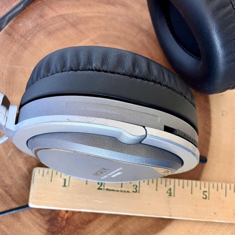 May include: Over-ear headphones with a silver and gray frame and black ear pads. The headphones have the brand name "SONY" printed on the ear cup. A wooden ruler is placed next to the headphones.