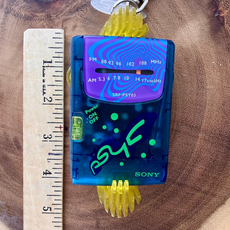May include: A blue and purple Sony SRF-PSY03 radio with a yellow plastic accent. The radio has a frequency dial and the text "Power ON OFF". A wooden ruler with inch measurements is next to the radio.