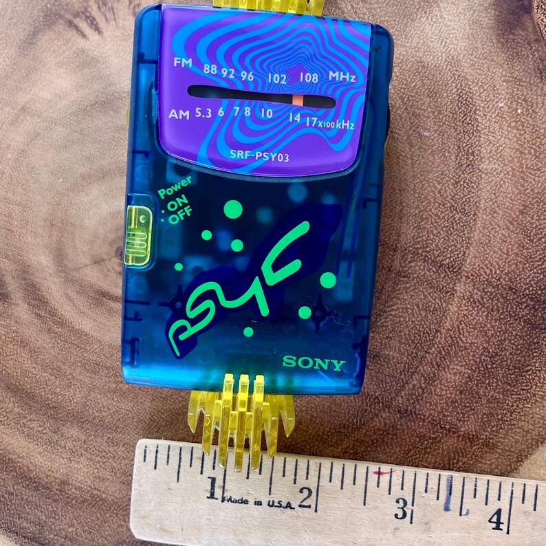 May include: A vintage blue Sony SRF-PSY03 radio with a purple and blue wave design. The radio has a power button, frequency dial, and the text "SONY". A yellow plastic piece is attached to the bottom. A ruler is next to the radio.