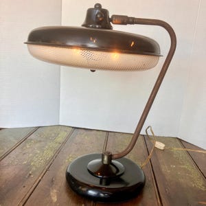 May include: Vintage desk lamp with a dark brown, oval shade and a perforated diffuser. The lamp has a curved, bronze-colored arm and a black, circular base. The lamp is plugged in and ready to use.