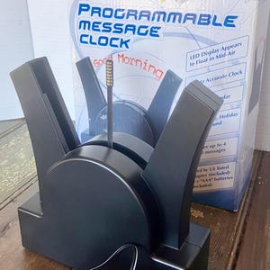 May include: A black programmable message clock with a unique design. The clock features a digital display that appears to float in mid-air and can display up to 4 messages. The box reads "Good Morning".