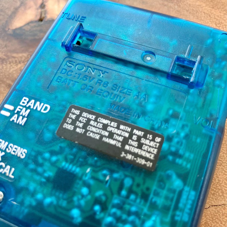 May include: A close-up of a transparent blue Sony radio. The radio has the text "TUNE", "BAND", "FM", "AM", and "EM SENS". The radio also has a sticker that says "THIS DEVICE COMPLIES WITH PART 15 OF THE FCC RULES..."