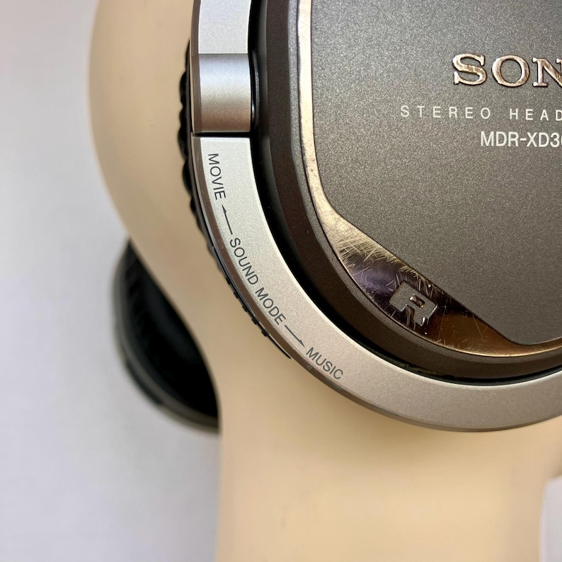 May include: Close-up of a gray Sony MDR-XD300 stereo headphone. The headphone features a silver trim with the text "SOUND MODE - MOVIE - MUSIC" and the Sony logo. The right side of the headphone is visible.