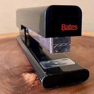 May include: A black Bates stapler with a silver metal base. The word "Bates" is printed in red on the top of the stapler. The stapler is on a wooden surface.