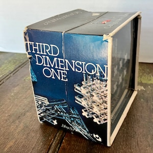 May include: A blue box with the words "Third Dimension One" printed on the front. The box contains a clear plastic puzzle with a geometric design. The box is made of cardboard and has a clear plastic window on the side.