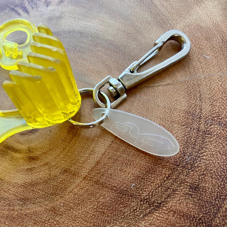 May include: A yellow, translucent keychain with a silver clasp and a small, oval tag. The keychain features a stylized hand design and a silver clasp for attaching to keys or a bag. The tag has a logo.