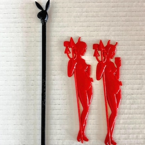 May include: A black swizzle stick featuring a rabbit head silhouette at the top and two red plastic stirrers, each shaped like a figure holding a trident and a glass, with a smooth finish and elongated stems for mixing beverages in glassware.