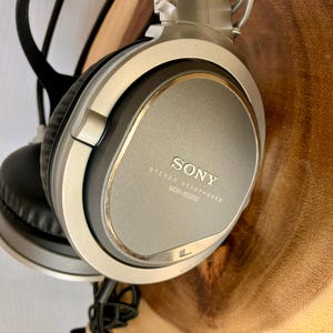 May include: Silver and black Sony MDR-XD300 stereo headphones. The headphones have large, round earcups and a padded headband. The Sony logo and model information are printed on the earcup. The headphones are shown against a wooden background.