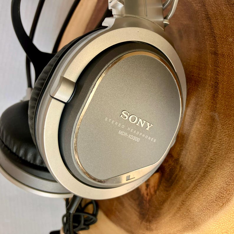 May include: Silver and black Sony MDR-XD300 stereo headphones. The headphones have large, round earcups and a padded headband. The Sony logo and model information are printed on the earcup. The headphones are shown against a wooden background.