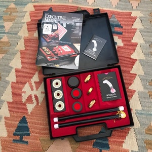 May include: A black carrying case with a red interior containing a magic kit. The kit includes two black magic wands, a set of red and black magic rings, a set of black and white magic rings, a set of red and black magic cups, and a deck of cards with the words "Wizard Magic Cards" printed on the front. The case also includes a booklet titled "The Executive Magic Collection" with the words "Secret Instructions" printed on the front.