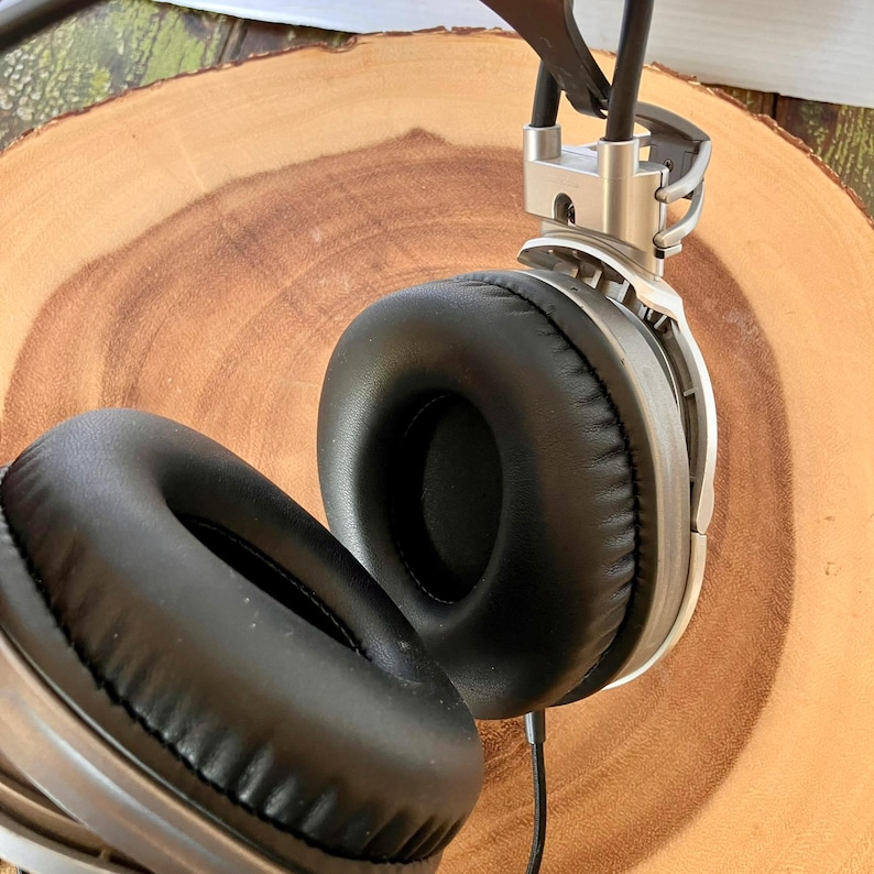 May include: Silver and black over-ear headphones with large, round earcups. The headphones have a silver metal frame and black padded earcups. A black cable extends from the headphones. The headphones are resting on a wooden surface.