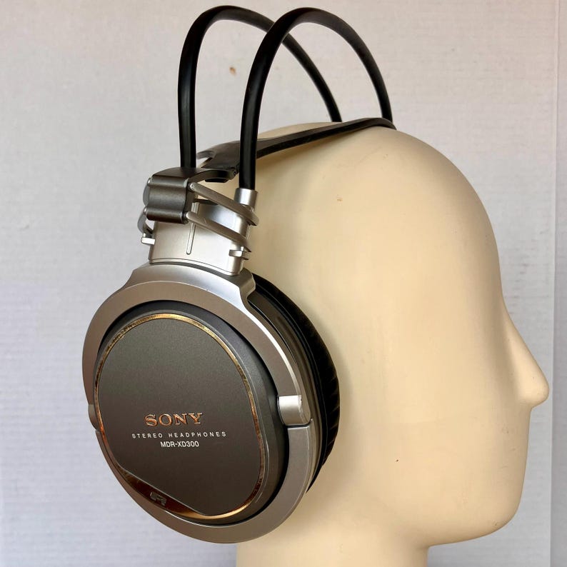 May include: Silver and gray Sony MDR-XD300 stereo headphones. The earcups are a dark gray with a gold-colored trim. The headband is black and the frame is silver. The headphones are on a mannequin head.