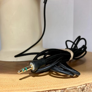 May include: A black audio cable with a gold-colored 3.5mm jack and green stripes. The cable is coiled and secured with a rubber band. The background includes a cream-colored object and a wooden surface.