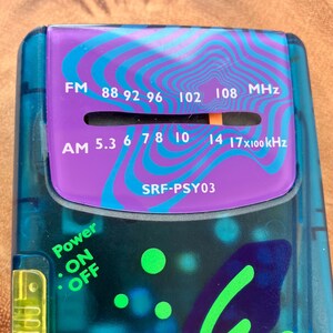 May include: A close-up of a vintage teal and purple radio. The radio features FM and AM frequency dials, with the text "FM 88 92 96 102 108 MHz" and "AM 5.3 6 7 8 10 14 17x100kHz". The radio also has a power button labeled "ON OFF" and the model number "SRF-PSY03".