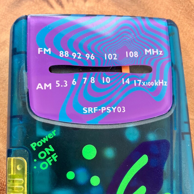 May include: A close-up of a vintage teal and purple radio. The radio features FM and AM frequency dials, with the text "FM 88 92 96 102 108 MHz" and "AM 5.3 6 7 8 10 14 17x100kHz". The radio also has a power button labeled "ON OFF" and the model number "SRF-PSY03".