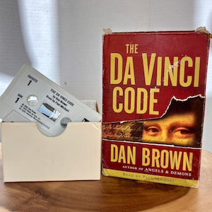 May include: A copy of Dan Brown's "The Da Vinci Code" book next to a cassette tape and a box of cassette tapes. The book has a red cover with yellow text and an image of eyes. The cassette tape is white with black text.