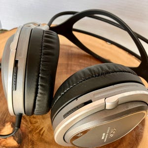 May include: Silver and black over-ear headphones with a black headband. The earcups are black and have a silver trim. The headphones have a volume control and a stereo recording label. The headphones are on a wooden surface.