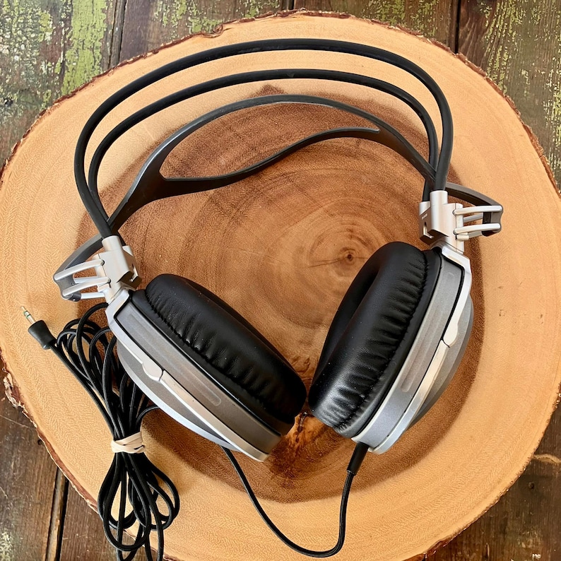 May include: Silver and black over-ear headphones with a black cord and a 3.5mm jack. The headphones have black earcups and a silver frame. The headphones are resting on a wooden surface.