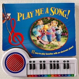 May include: A children's musical toy featuring a white electronic piano with black and white keys and a red speaker. The toy is paired with a blue book titled "PLAY ME A SONG!" with illustrations of children dancing. The book includes the text "12 easy-to-play favorites with an electronic piano!"