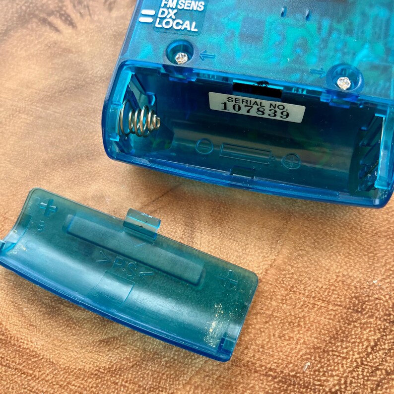 May include: A transparent blue vintage radio with an open battery compartment. The serial number 107839 is visible on a sticker. The radio's control panel includes the text "FM SENS DX LOCAL".