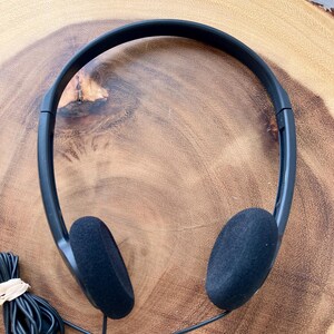 May include: Black over-the-ear headphones with foam ear pads. The headphones have a black headband and a black cord. The cord is bundled with a rubber band. The headphones are on a wooden surface.