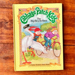 May include: A hardcover children's book titled Cabbage Patch Kids The Big Bicycle Race. The yellow cover features a large white stork wearing a hat, a child in a red cap on a bicycle, and a child with blonde hair on a red bicycle in a park.