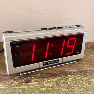 May include: A silver digital alarm clock with a red LED display showing the time 11:19 PM. The clock has a "Micronta" brand name on the front.