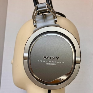 May include: Silver Sony MDR-XD300 stereo headphones. The headphones have a circular ear cup design with the Sony logo and text. The headband and ear cup frames are silver. The headphones are on a mannequin head.