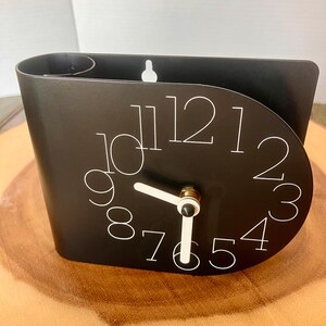 May include: A black wall clock with white numerals and hands. The clock has a unique curved design and a small hook for hanging. The clock face is a semi-circle shape. The clock is sitting on a wooden surface.