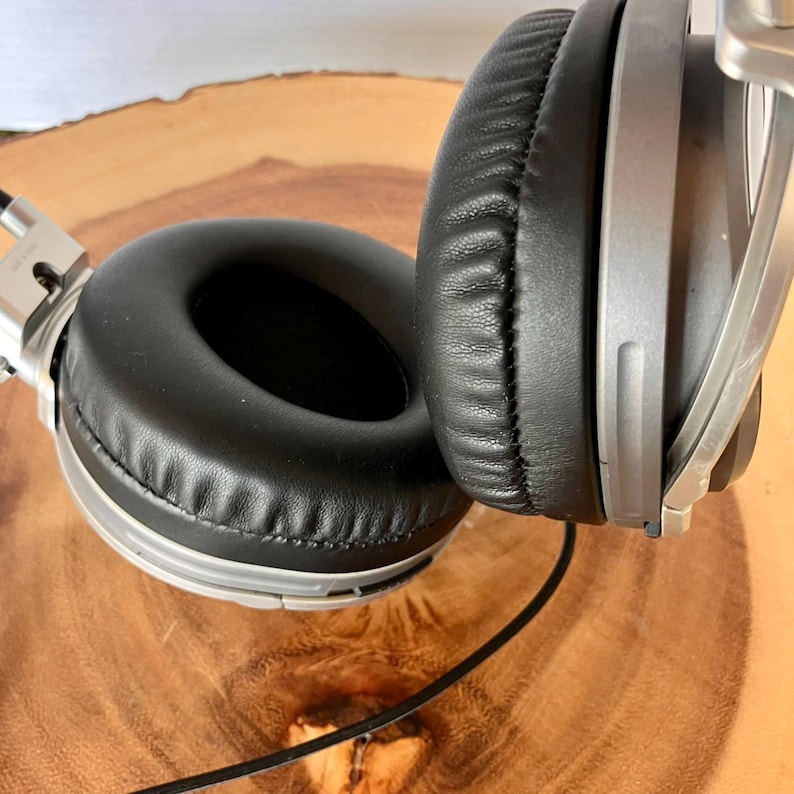 May include: Silver and black over-ear headphones with a padded black ear cup and a silver frame. The headphones are shown on a wooden surface. A black cord extends from the headphones.