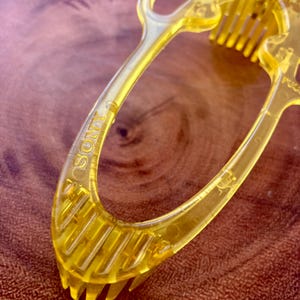 May include: Close-up of a transparent yellow plastic hair comb with the word "SONY" printed on the handle. The comb has multiple teeth and a curved handle. The comb is on a brown wooden surface.