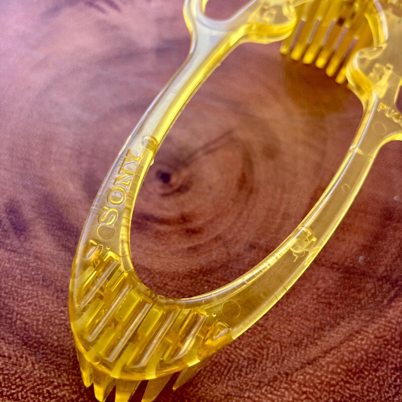 May include: Close-up of a transparent yellow plastic hair comb with the word "SONY" printed on the handle. The comb has multiple teeth and a curved handle. The comb is on a brown wooden surface.