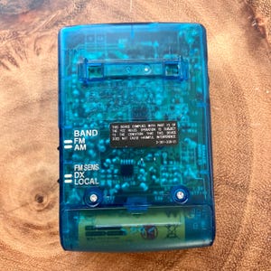 May include: A transparent blue Sony radio with visible internal components. The radio has buttons for band selection, FM sensitivity, and local/DX. Text on the device includes "SONY" and regulatory information. A battery is visible at the bottom.
