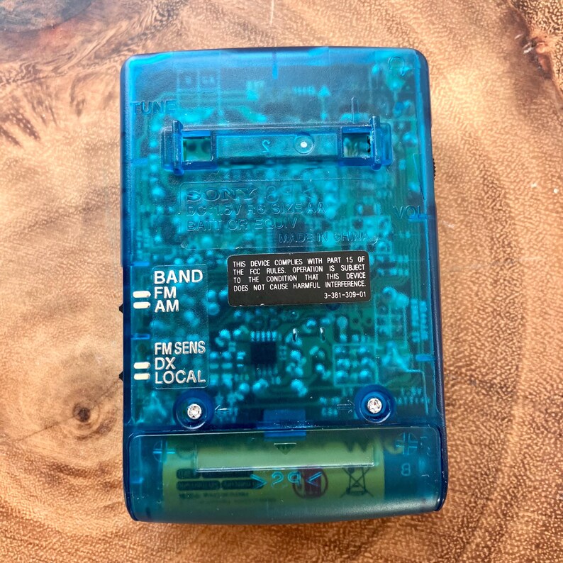 May include: A transparent blue Sony radio with visible internal components. The radio has buttons for band selection, FM sensitivity, and local/DX. Text on the device includes "SONY" and regulatory information. A battery is visible at the bottom.