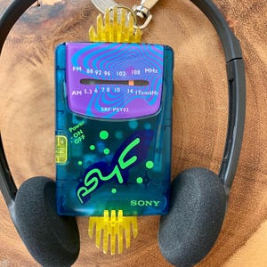 May include: A vintage Sony SRF-PSY03 Walkman radio with a blue and purple design, featuring FM and AM radio frequencies. The radio has a power button and the word "PSY" in green. Black headphones are attached.