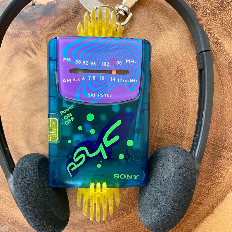 May include: A vintage Sony SRF-PSY03 Walkman radio with a blue and purple design, featuring FM and AM radio frequencies. The radio has a power button and the word "PSY" in green. Black headphones are attached.