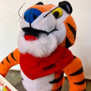 May include: A plush orange and black tiger wearing a red bandana with the text "TONY" on it. The tiger has a white mustache and a blue nose.