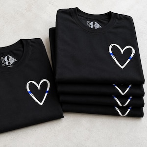 May include: A stack of black t-shirts and one single black t-shirt laid flat. Each shirt features a white hand-drawn heart outline on the left chest, accented with a thin horizontal blue line. The inner neck label displays the text 9 FLOOR.