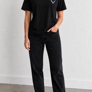 May include: A black short-sleeved t-shirt featuring a small white heart graphic with a thin blue horizontal line detail on the left chest, paired with black denim jeans and white sneakers. The t-shirt has a crew neckline and a standard fit.