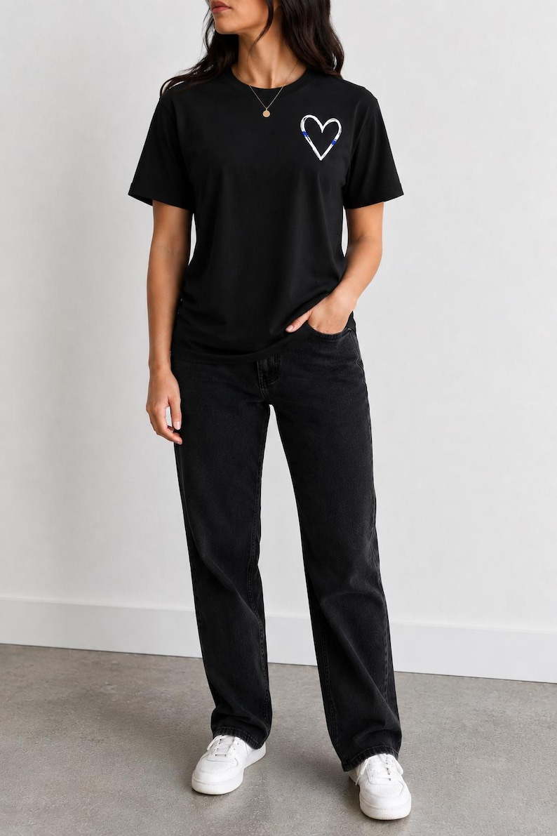 May include: A black short-sleeved t-shirt featuring a small white heart graphic with a thin blue horizontal line detail on the left chest, paired with black denim jeans and white sneakers. The t-shirt has a crew neckline and a standard fit.