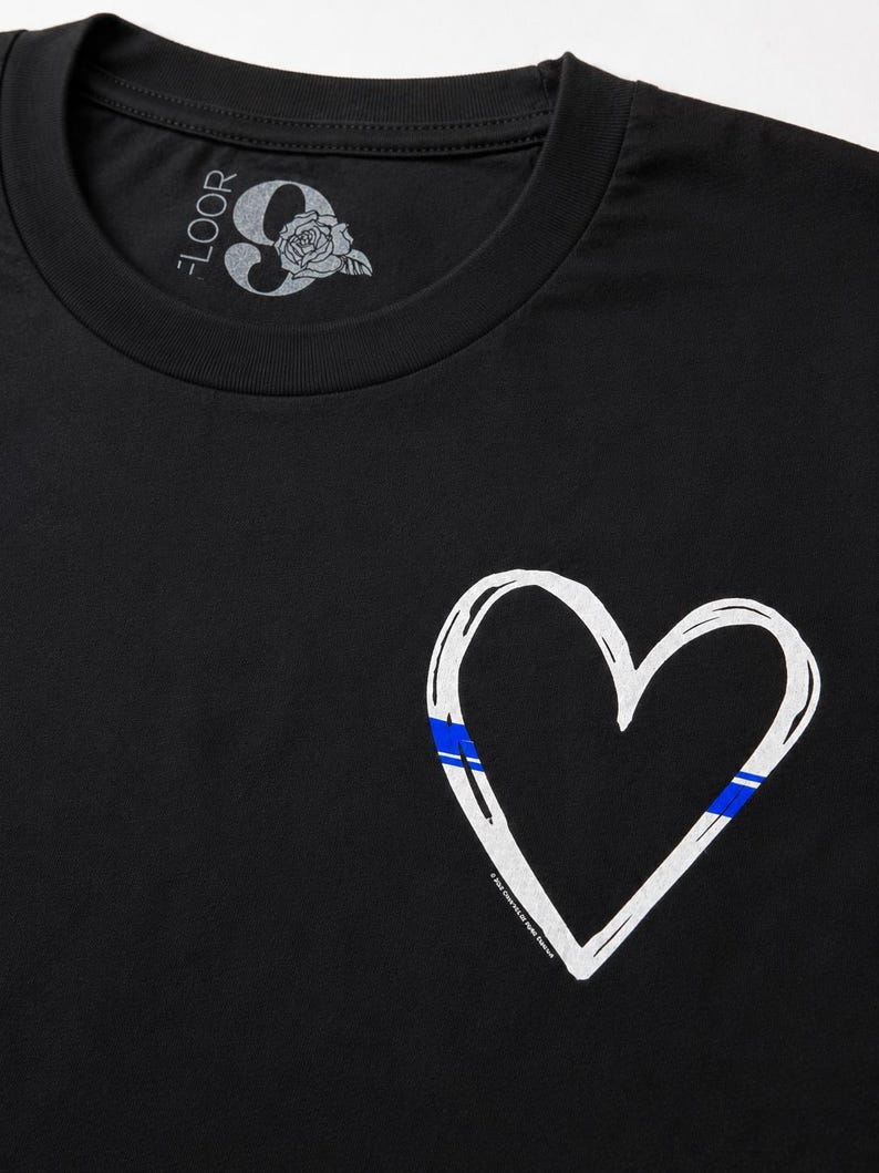 May include: A black t-shirt featuring a white hand-drawn heart outline on the front. The heart includes a thin blue horizontal stripe detail. The inner neck label displays the text 9 FLOOR with a rose graphic and additional small text.