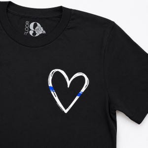 May include: A black short-sleeved t-shirt featuring a white hand-drawn heart graphic on the chest. The heart outline includes a thin horizontal blue stripe. The inner neck label displays the text FLOOR 9 with a rose illustration above the number.