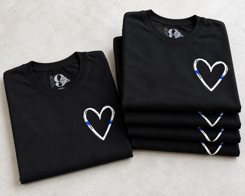 May include: A stack of black t-shirts and one single black t-shirt laid flat. Each shirt features a white hand-drawn heart outline on the left chest, accented with a thin horizontal blue line. The inner neck label displays the text 9 FLOOR.