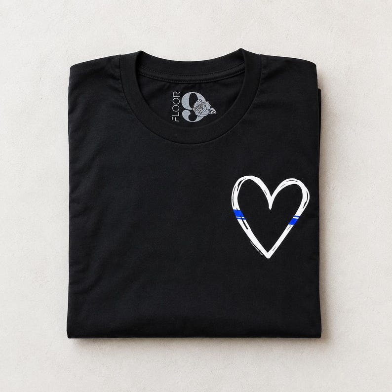 May include: A black short-sleeved t-shirt featuring a white hand-drawn heart outline on the front right side. The heart includes a thin horizontal blue stripe. The inner neck label displays the text FLOOR 9 with a rose graphic illustration.