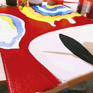 May include: A close-up of a canvas painting in progress. The painting features abstract shapes in red, white, blue, yellow, and black. A hand is holding a paintbrush and applying black paint to the canvas.
